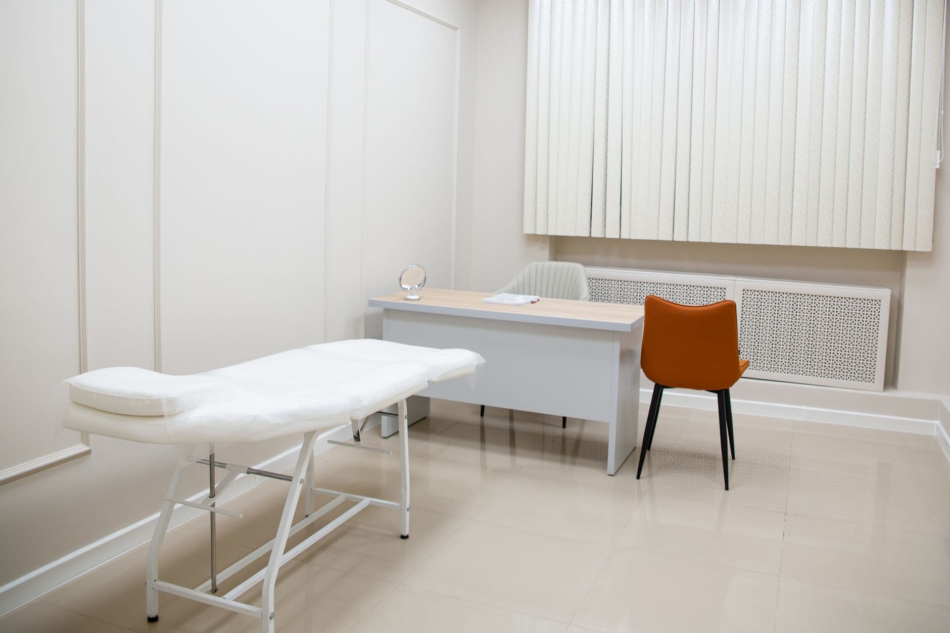 A minimalist medical examination room featuring a desk, chairs, and an examination bed in a sterile, neutral s